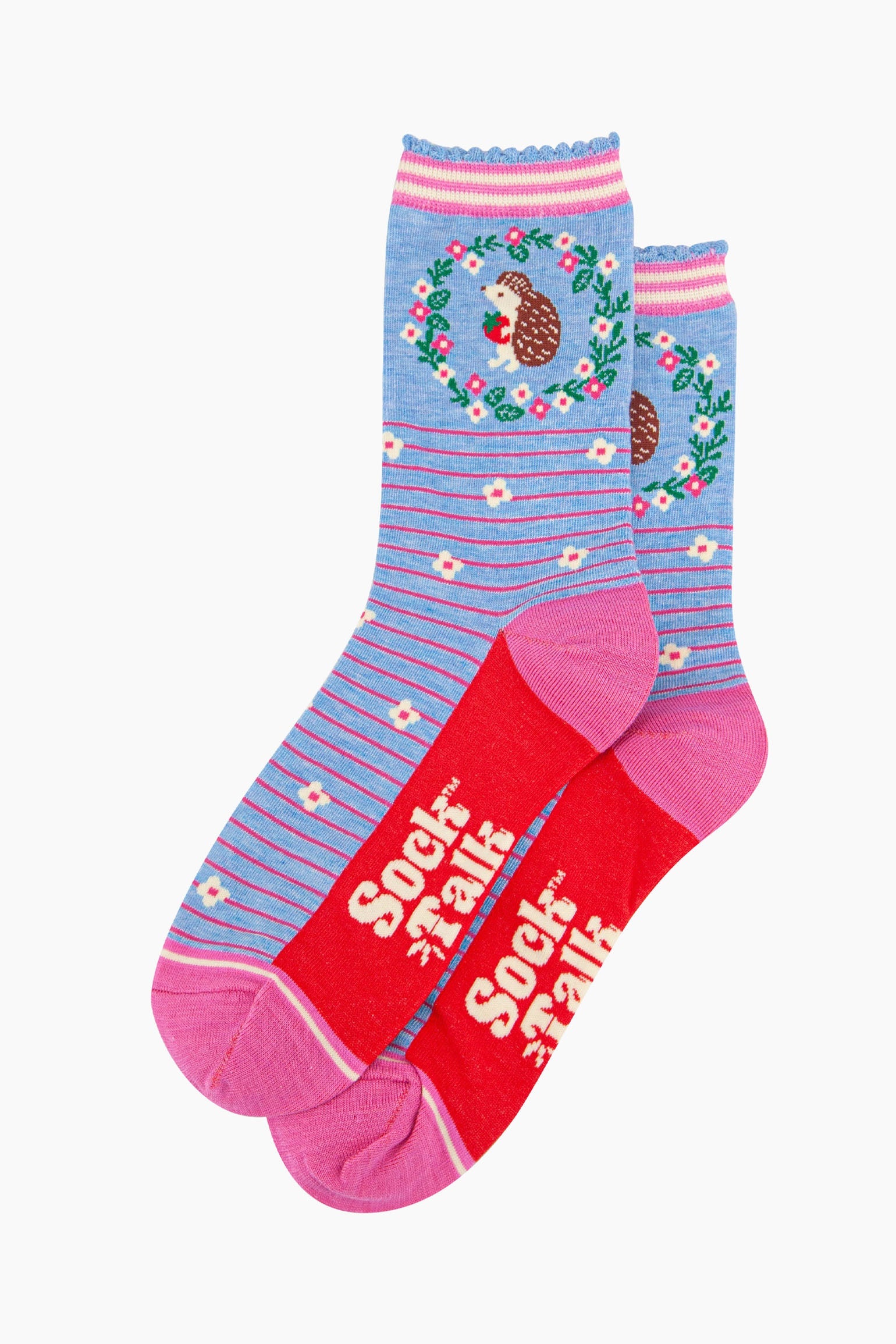 the pair of blue hedgehog socks laying flat one overlapping the other. showing clearly the pastel blue background of the socks which is contrasted by pink pinstripes and scattered white flowers. on the ankle of the socks there is a woodland hedgehog holding a strawberry, the hedgehog is surrounded by a pink, white and green floral wreath the sock talk logo is visible on the base of the sole.