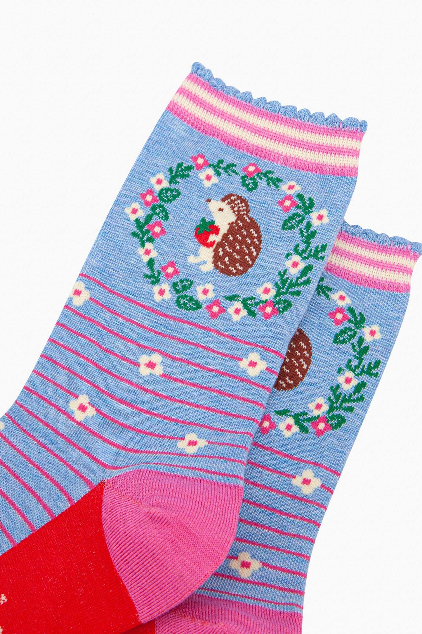 close up of the pattern. on the ankle of the socks is a large brown and beige hedgehog standing on its hind legs. it is holding a red strawberry in its hands. surrounding the hedgehog is a floral wreath made of white flowers, pink flowers and green leaves.