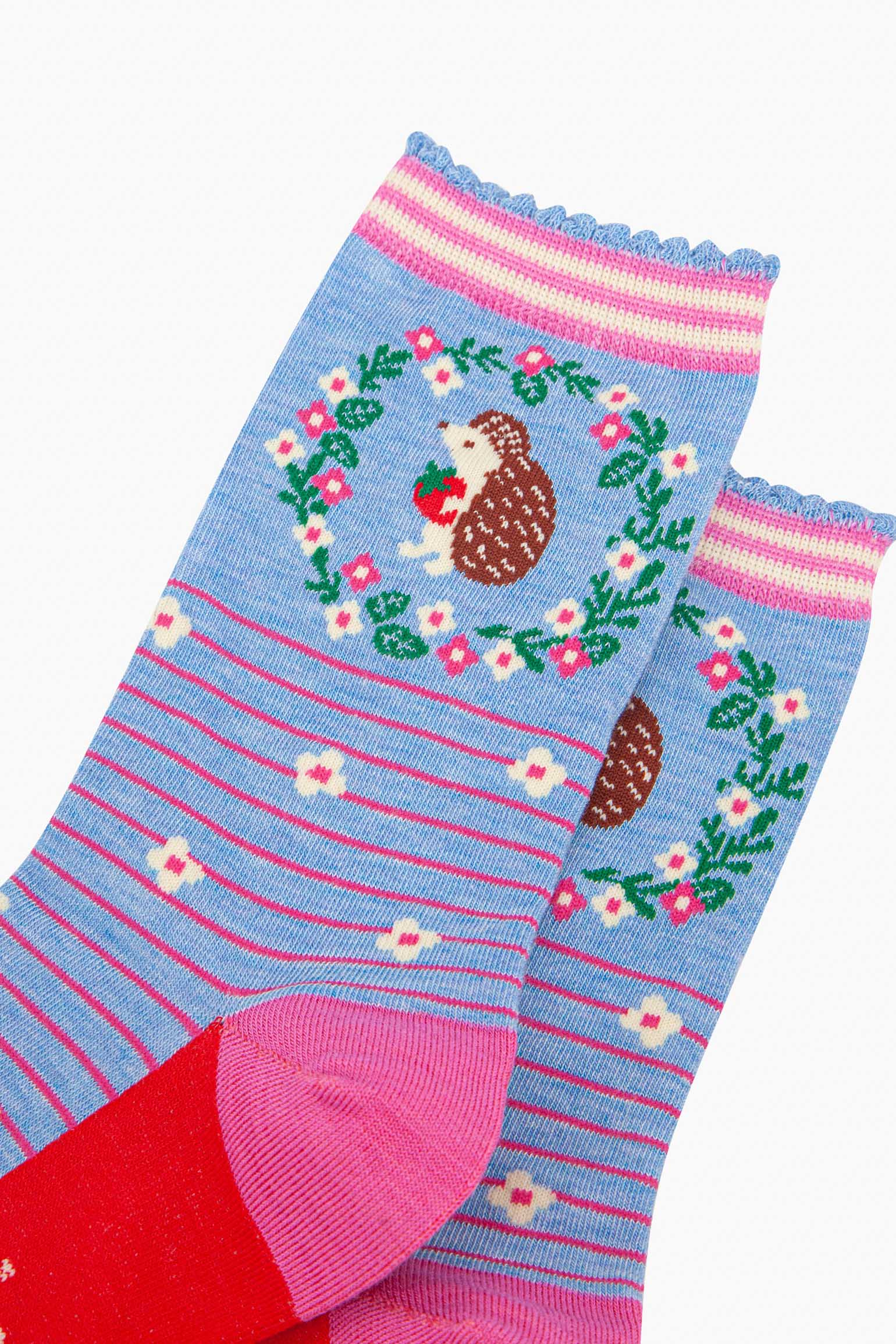 close up of the pattern. on the ankle of the socks is a large brown and beige hedgehog standing on its hind legs. it is holding a red strawberry in its hands. surrounding the hedgehog is a floral wreath made of white flowers, pink flowers and green leaves.