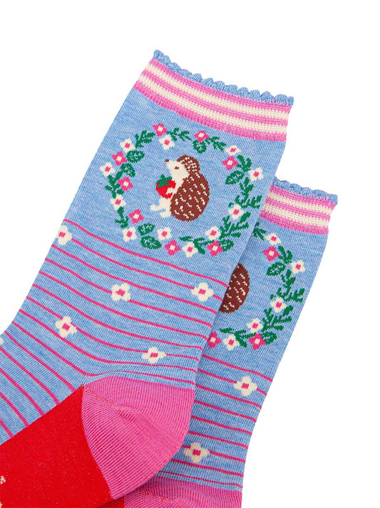 close up of the pattern. on the ankle of the socks is a large brown and beige hedgehog standing on its hind legs. it is holding a red strawberry in its hands. surrounding the hedgehog is a floral wreath made of white flowers, pink flowers and green leaves.