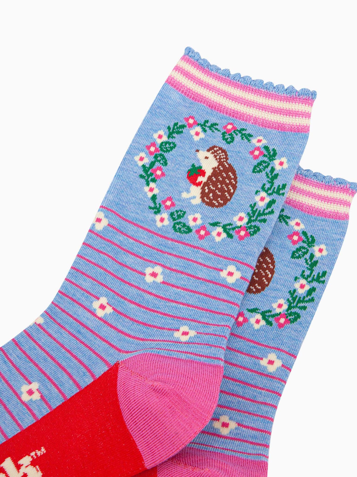 close up of the pattern. on the ankle of the socks is a large brown and beige hedgehog standing on its hind legs. it is holding a red strawberry in its hands. surrounding the hedgehog is a floral wreath made of white flowers, pink flowers and green leaves.