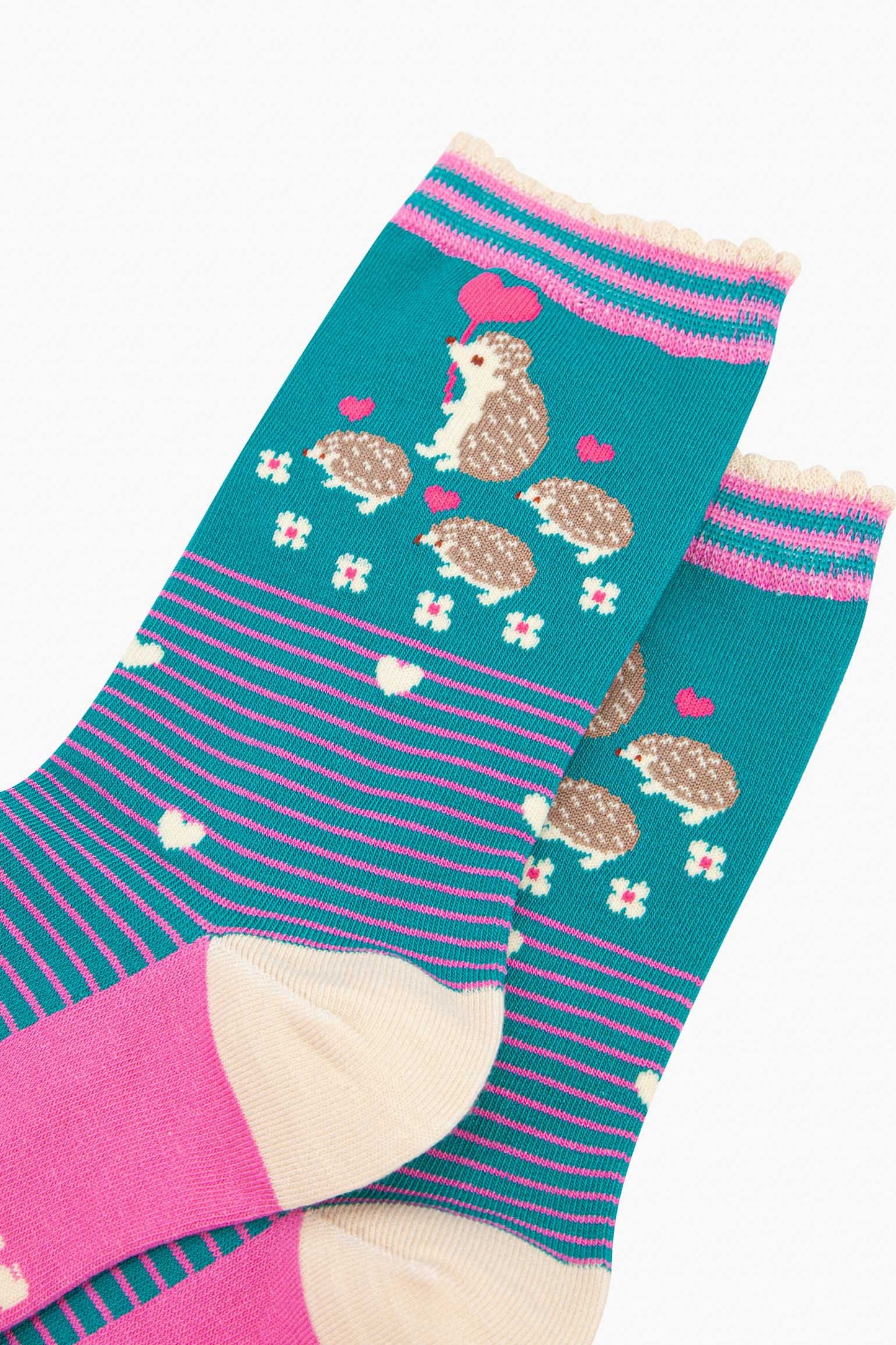 close up of the pattern. there are four hedgehogs on the ankle, one adult and three babies they are beige and each are carrying a fuchsia pink love heart shaped balloon. there are six small white flowers in a semi circle around the bottom of the family. the body of the socks has pink pinstripes and scattered white love hearts.