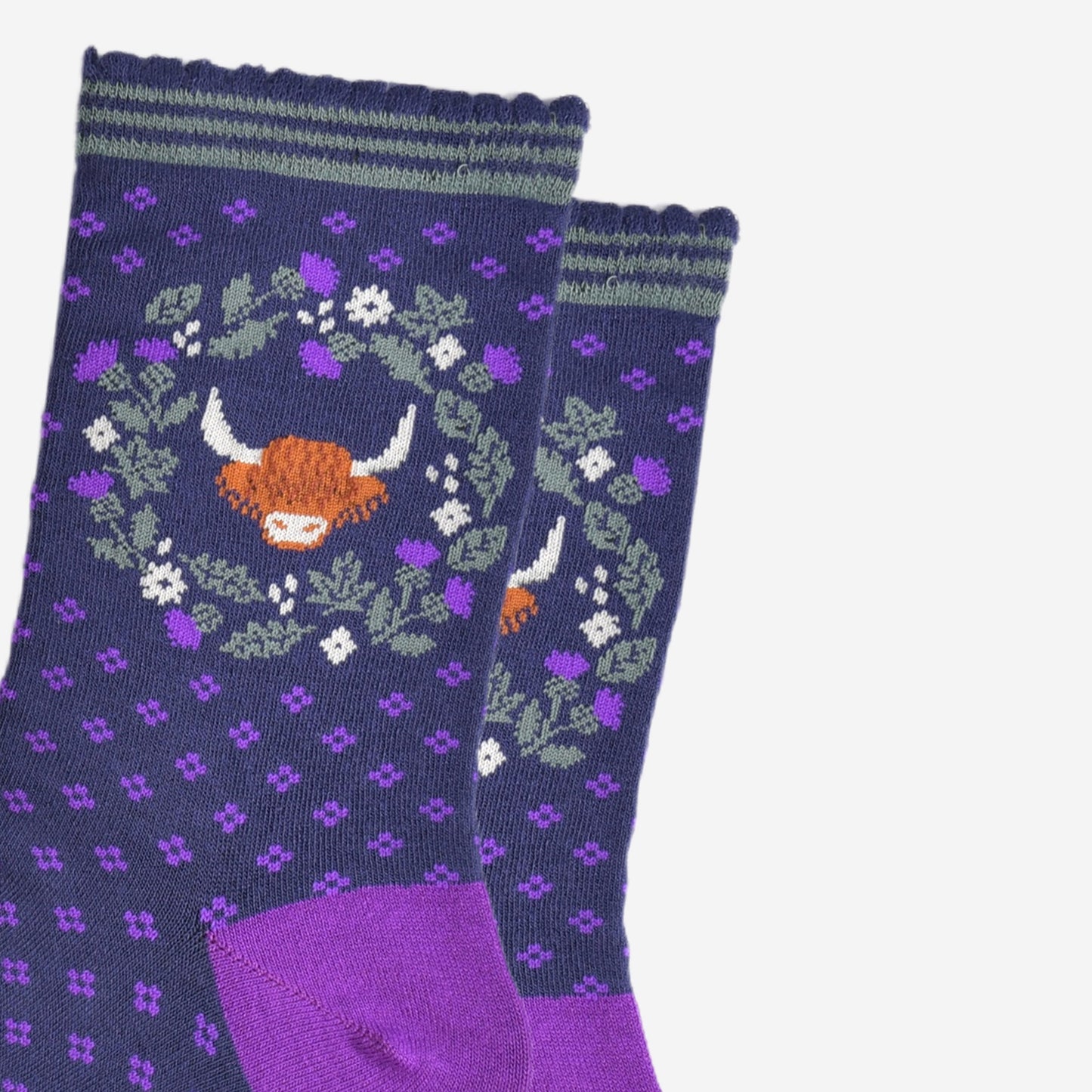 close up of the highland cow and wreath pattern on the ankle of the socks. the wreath is made from green and purple scottish thistles, the cow's head is in the middle of the wreath, the cow has long shaggy hair covering its eyes