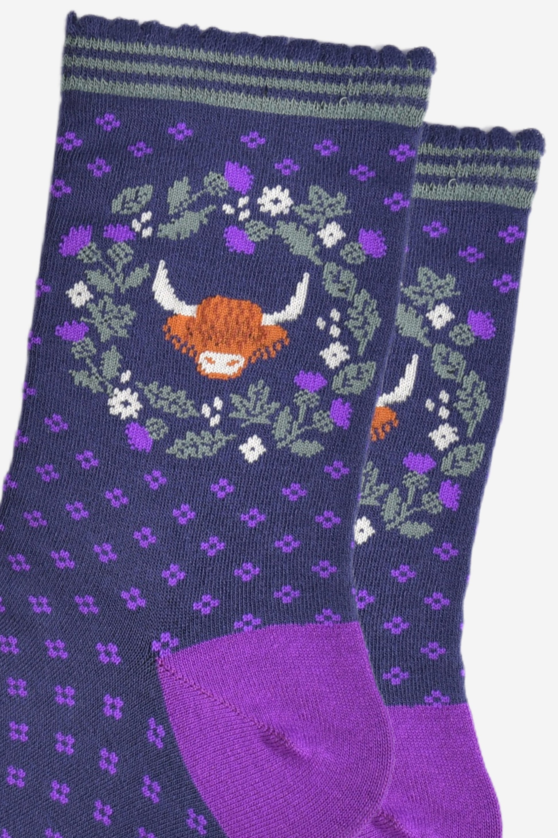 close up of the highland cow and wreath pattern on the ankle of the socks. the wreath is made from green and purple scottish thistles, the cow's head is in the middle of the wreath, the cow has long shaggy hair covering its eyes