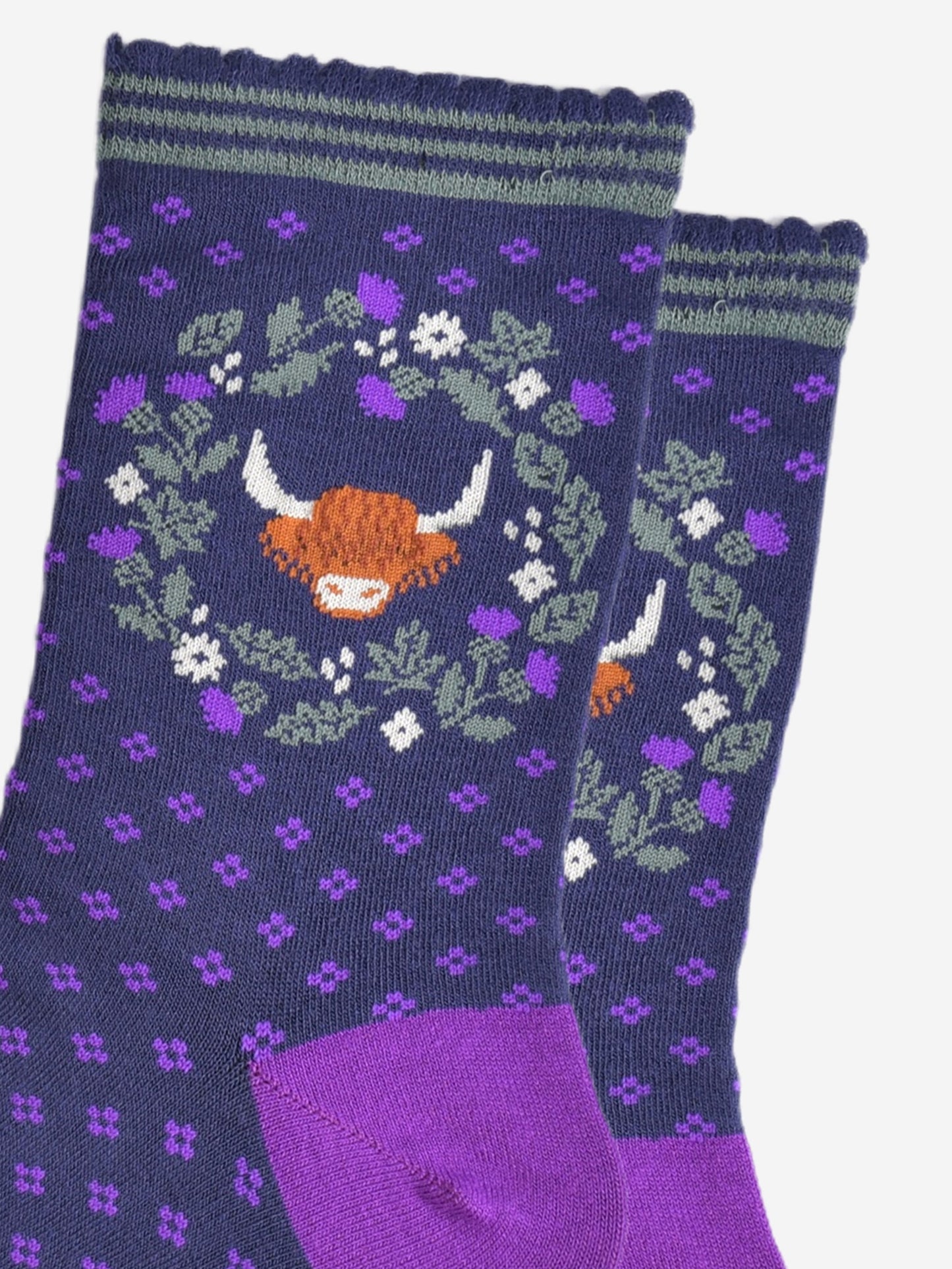 close up of the highland cow and wreath pattern on the ankle of the socks. the wreath is made from green and purple scottish thistles, the cow's head is in the middle of the wreath, the cow has long shaggy hair covering its eyes