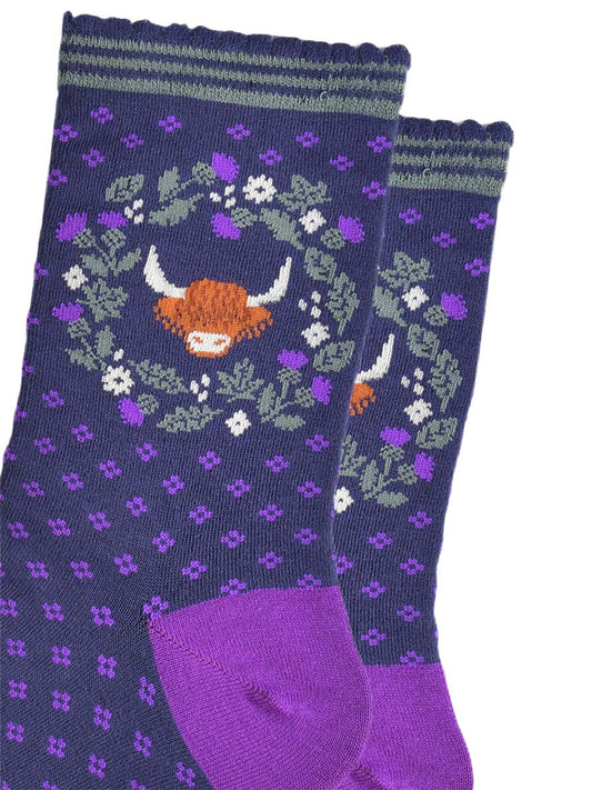 close up of the highland cow and wreath pattern on the ankle of the socks. the wreath is made from green and purple scottish thistles, the cow's head is in the middle of the wreath, the cow has long shaggy hair covering its eyes