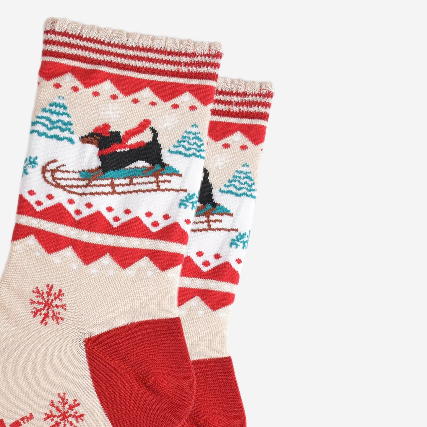 close up of the sledging sausage dog pattern, the dog is a traditional black dachshund, it is standing on a sledge and wearing a red winter hat and scarf, behind the dog are snow covered trees