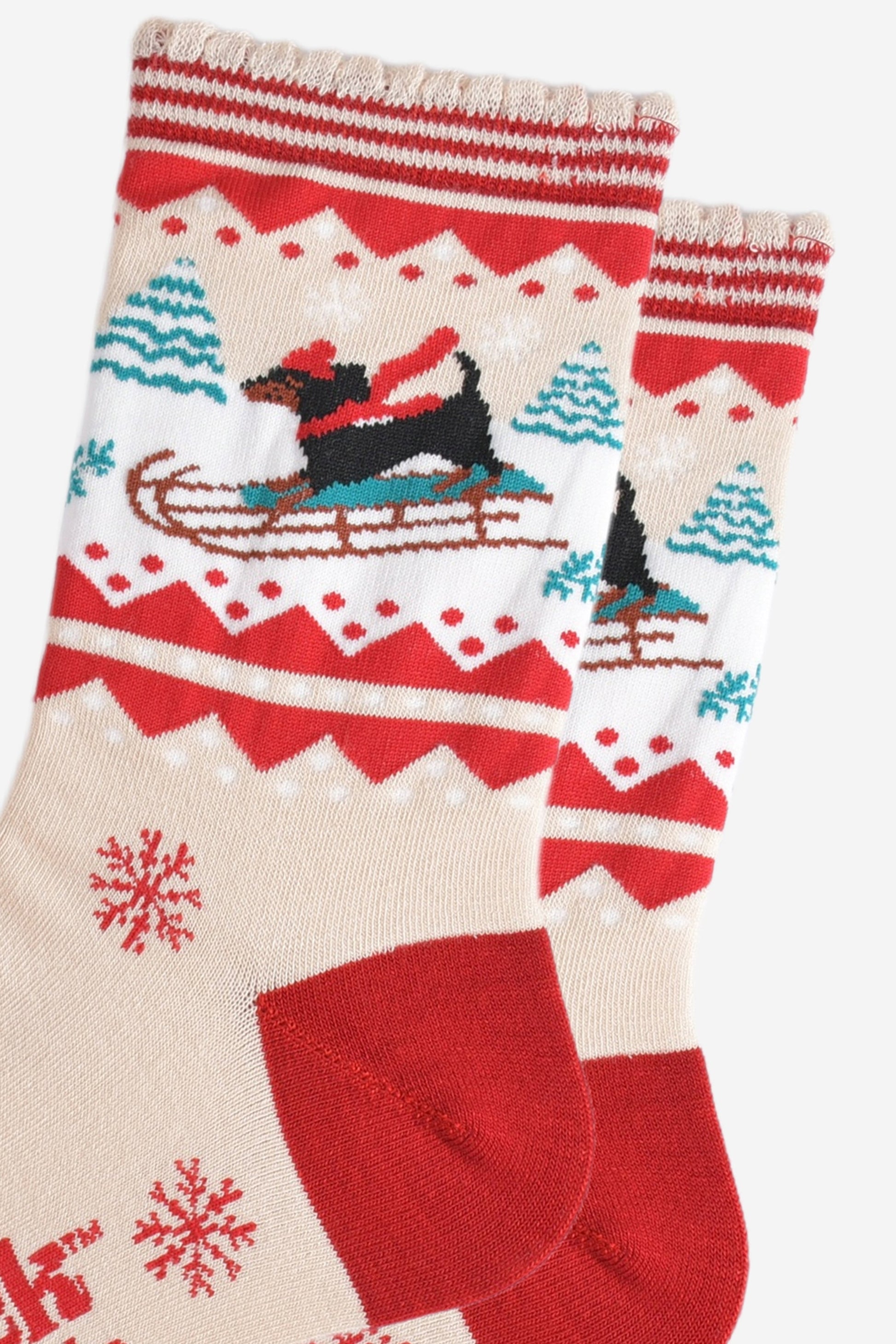 close up of the sledging sausage dog pattern, the dog is a traditional black dachshund, it is standing on a sledge and wearing a red winter hat and scarf, behind the dog are snow covered trees
