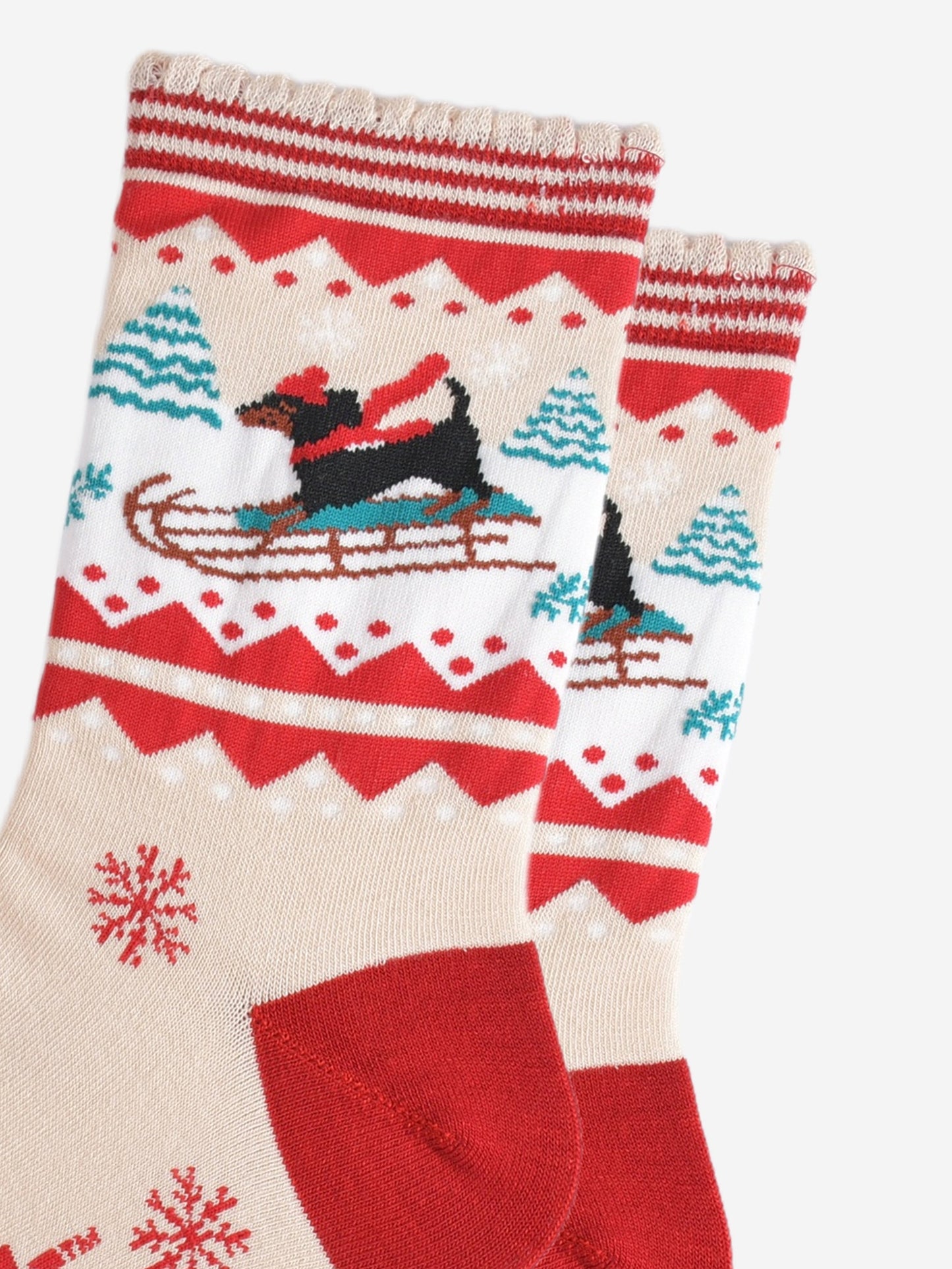 close up of the sledging sausage dog pattern, the dog is a traditional black dachshund, it is standing on a sledge and wearing a red winter hat and scarf, behind the dog are snow covered trees