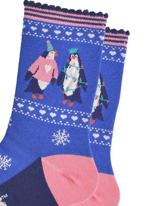 close up of the penguins on the ankle of the socks, one is wearing a pink xmas jumper and green party hat. the other penguin is entangled in festive party lights and is also wearing a crown shaped party hat