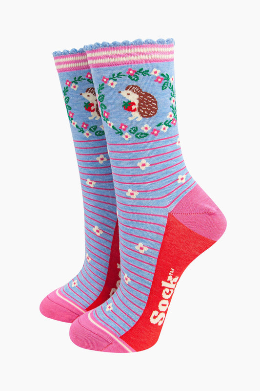 light blue bamboo socks with pink horizontal pinstripes. on the ankle of the socks there is a hedgehog holding a strawberry. around the hedgehog is a pink and white floral wreath.