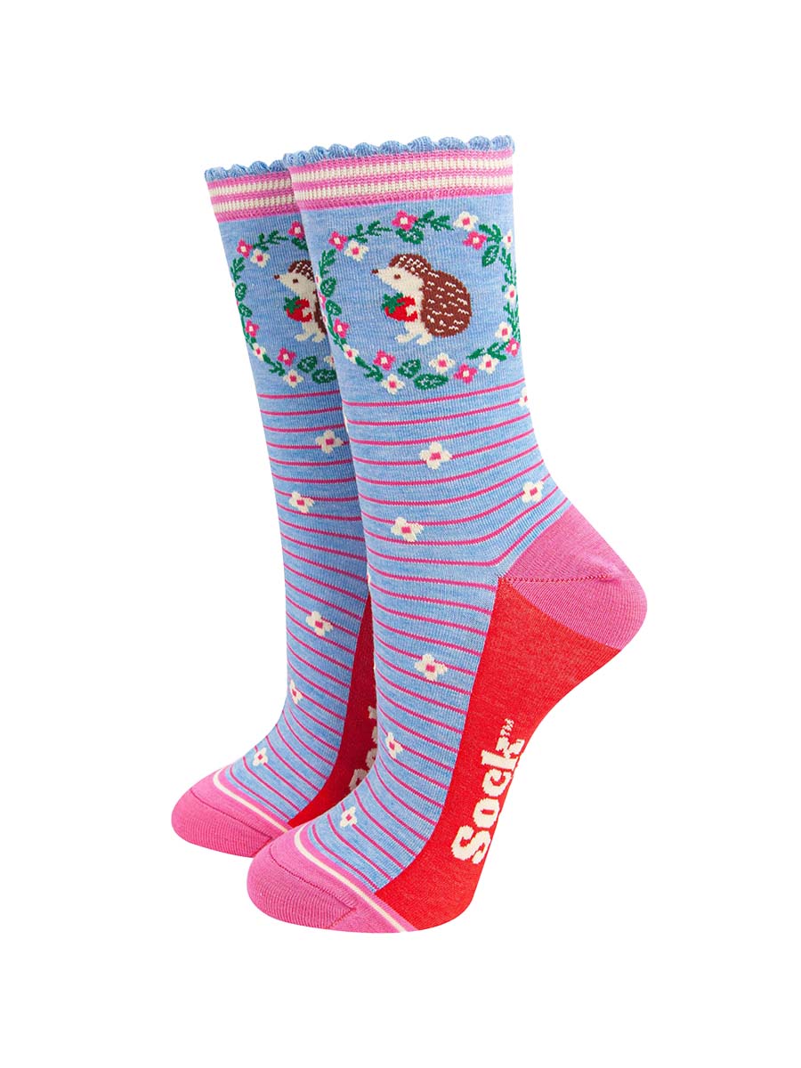 light blue bamboo socks with pink horizontal pinstripes. on the ankle of the socks there is a hedgehog holding a strawberry. around the hedgehog is a pink and white floral wreath.
