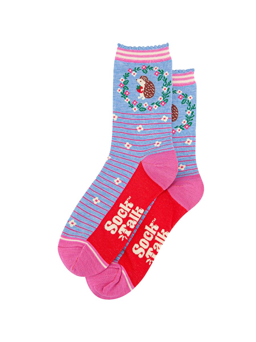 the pair of blue hedgehog socks laying flat one overlapping the other. showing clearly the pastel blue background of the socks which is contrasted by pink pinstripes and scattered white flowers. on the ankle of the socks there is a woodland hedgehog holding a strawberry, the hedgehog is surrounded by a pink, white and green floral wreath the sock talk logo is visible on the base of the sole. 
