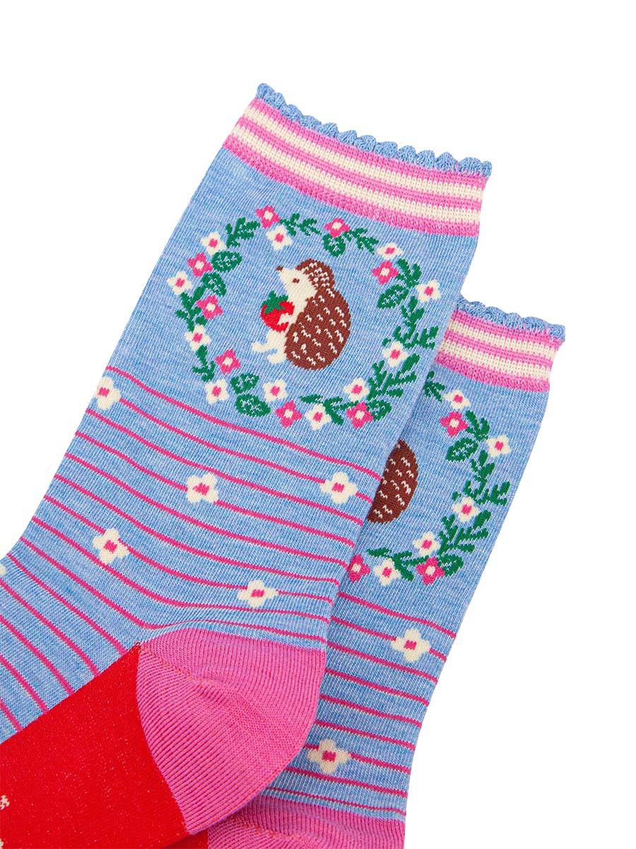 close up of the pattern. on the ankle of the socks is a large brown and beige hedgehog standing on its hind legs. it is holding a red strawberry in its hands. surrounding the hedgehog is a floral wreath made of white flowers, pink flowers and green leaves. 