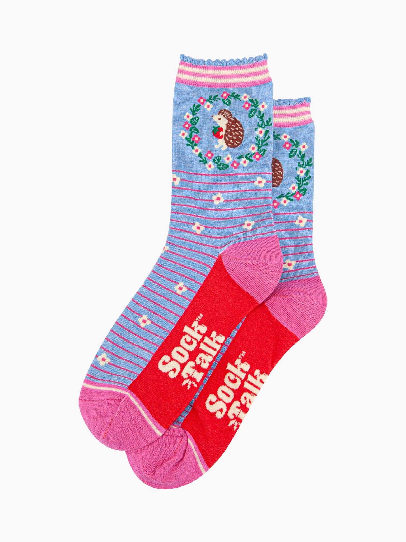 the pair of blue hedgehog socks laying flat one overlapping the other. showing clearly the pastel blue background of the socks which is contrasted by pink pinstripes and scattered white flowers. on the ankle of the socks there is a woodland hedgehog holding a strawberry, the hedgehog is surrounded by a pink, white and green floral wreath the sock talk logo is visible on the base of the sole. 