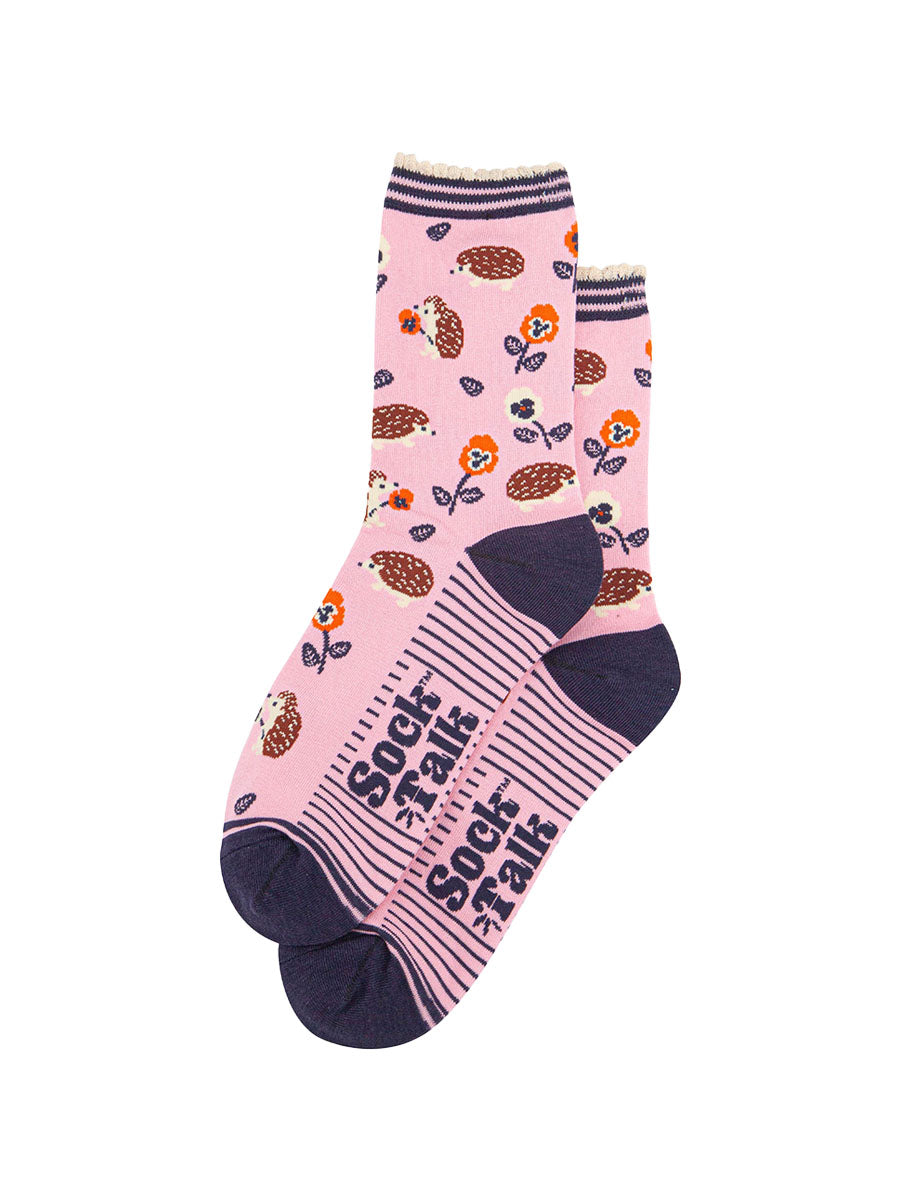 the pair of light pink hedgehog socks laying flat one overlapping the other. showing clearly the pink background of the socks which is contrasted by the all over pattern of hedgehogs, flowers and leaves.  the sock talk logo is navy blue and visible on the base of the sole.
