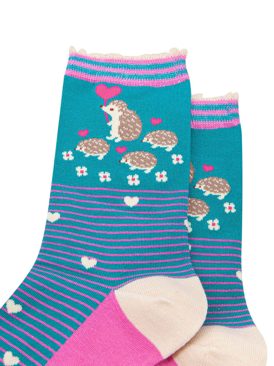 close up of the pattern. there are four hedgehogs on the ankle, one adult and three babies they are beige and each are carrying a fuchsia pink love heart shaped balloon. there are six small white flowers in a semi circle around the bottom of the family. the body of the socks has pink pinstripes and scattered white love hearts.