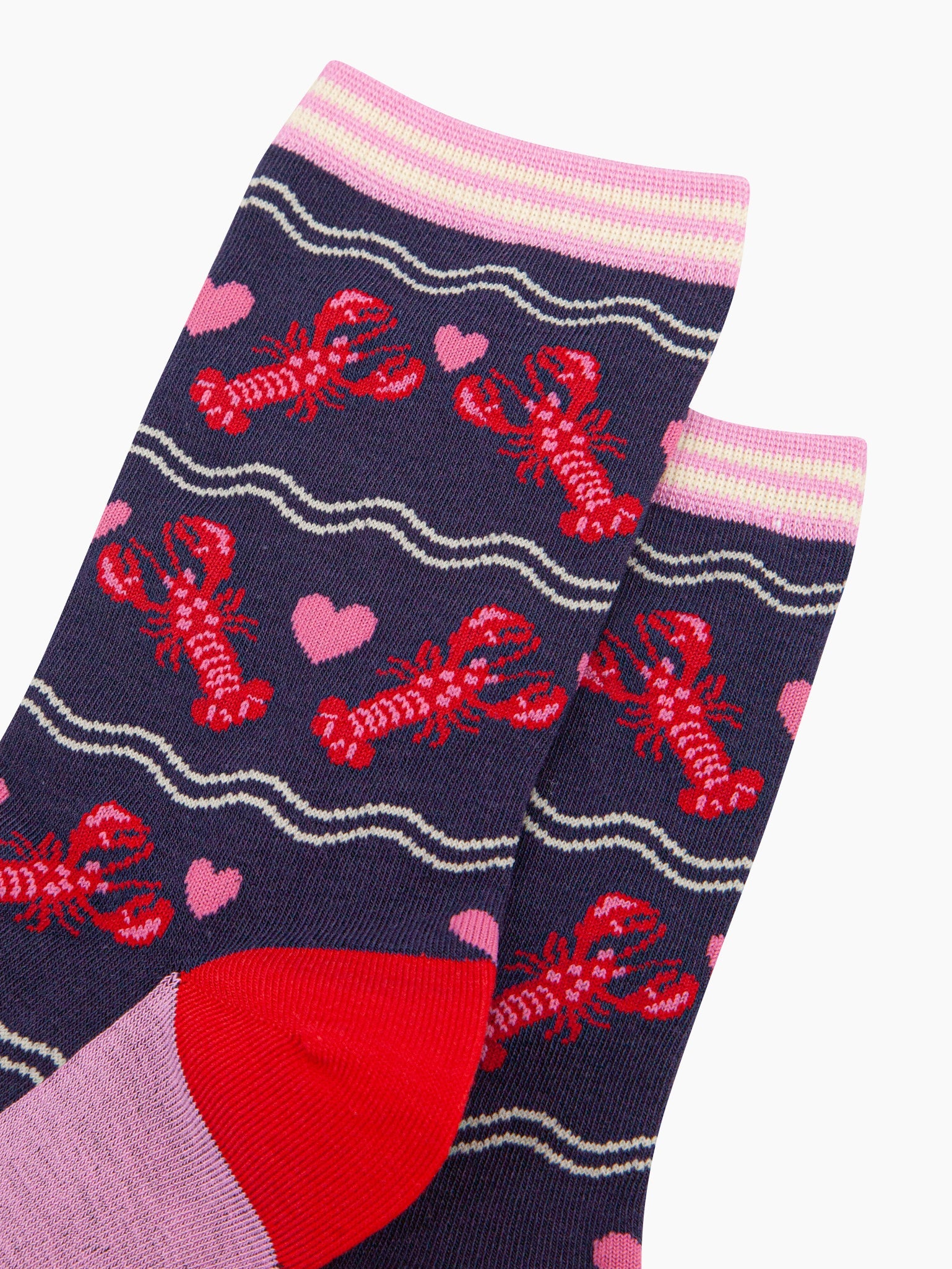 close up of the pattern, the lobsters are red with pink highlights on their backs and pincers, they are in pairs with a pink love heart between them. the cuff of the socks is straight cut and is light pink and cream striped.