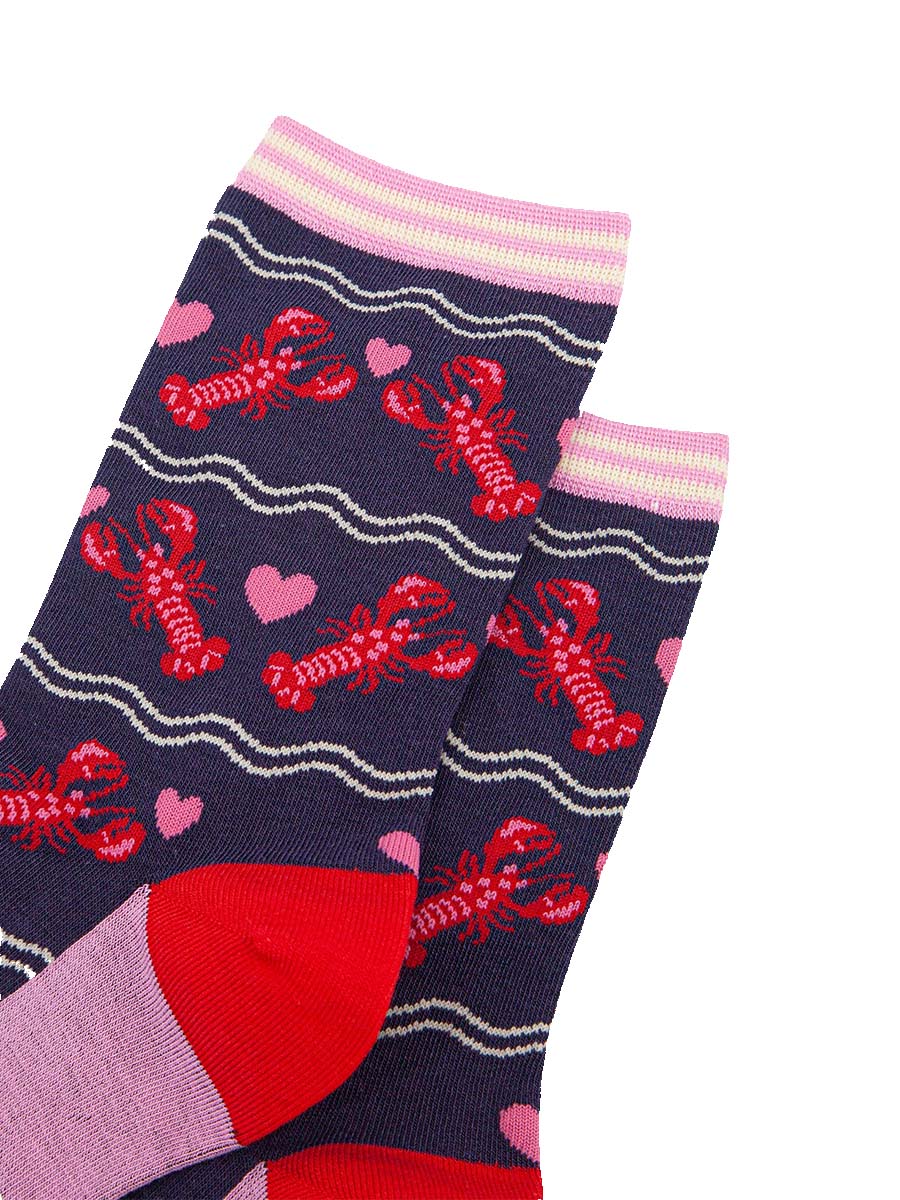 close up of the pattern, the lobsters are red with pink highlights on their backs and pincers, they are in pairs with a pink love heart between them. the cuff of the socks is straight cut and is light pink and cream striped.