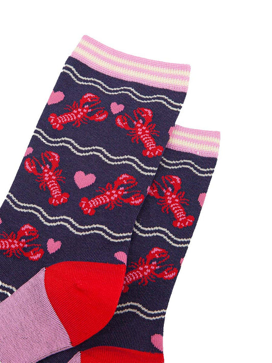 close up of the pattern, the lobsters are red with pink highlights on their backs and pincers, they are in pairs with a pink love heart between them. the cuff of the socks is straight cut and is light pink and cream striped.