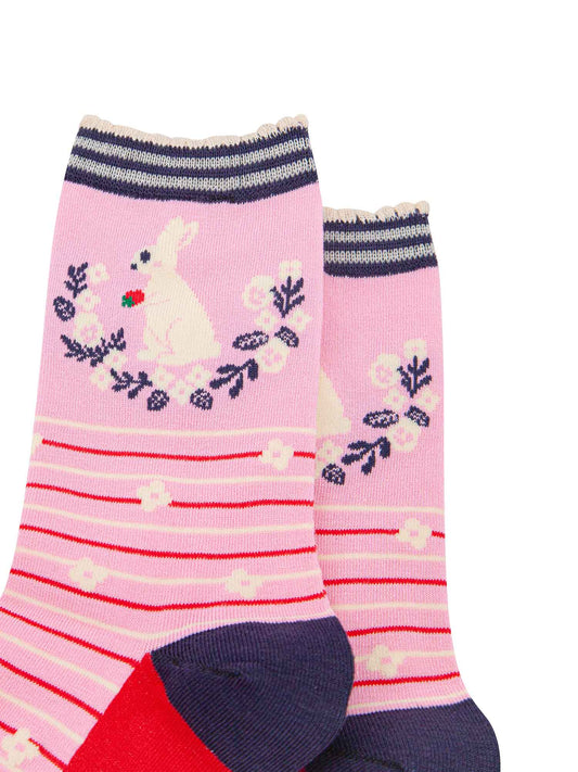 close up of the pattern. on the ankle of the socks is a large white rabbit sitting on its hind legs. it is holding a red strawberry in its paws. underneath the rabbit is a semi circular floral wreath made of white flowers and navy blue leaves.
