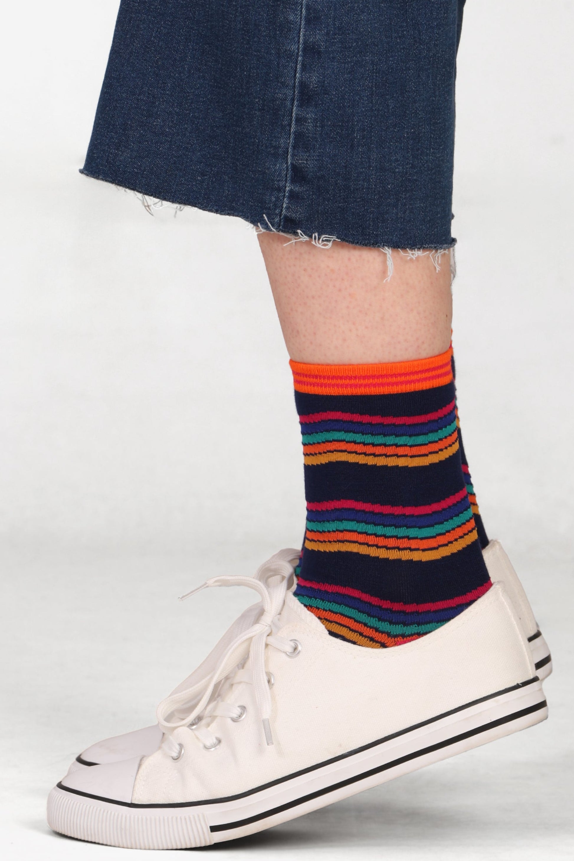 Model wearing rainbow stripe bamboo socks in a navy blue and multicolour design with cropped jeans and white trainers