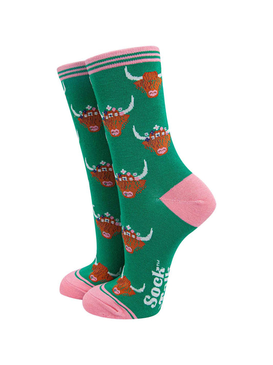 green ankle socks with pink toe, heel and cuff featuring a pattern of brown highland cow faces wearing multicoloured floral wreaths