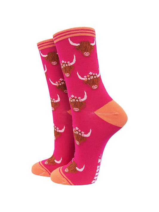 a bright pink ankle sock with a pattern of brown highland cow faces wearing floral crowns all over.