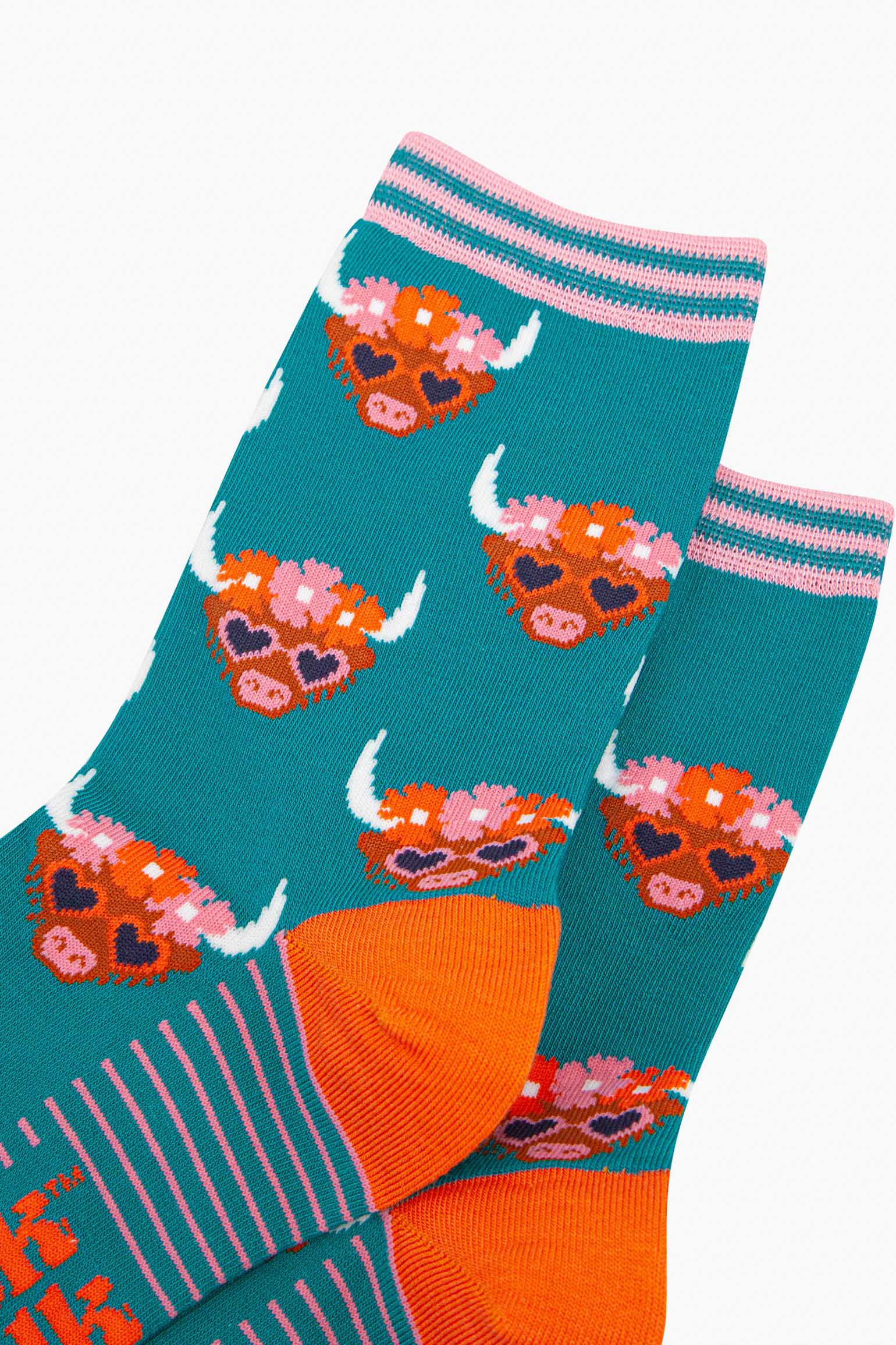 close up of the pattern. the highland cow faces are brown, they have white pointed horns and wear pink love heart sunglasses. on the heads of the cows is a floral wreath composed of pink and orange flowers. the cuff is teal and light pink striped.