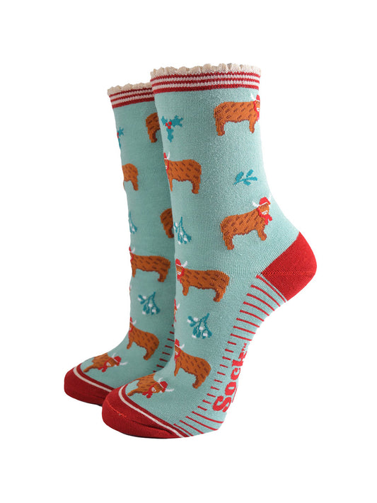 mint green bamboo socks with a red striped scalloped cuff and an all over pattern of highland cows wearing santa hats, the socks are further decorated with holly and mistletoe accents