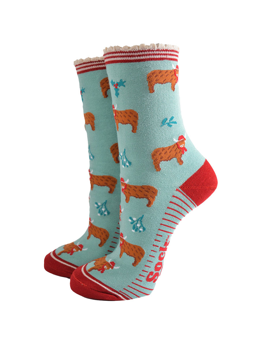 mint green bamboo socks with a red striped scalloped cuff and an all over pattern of highland cows wearing santa hats, the socks are further decorated with holly and mistletoe accents