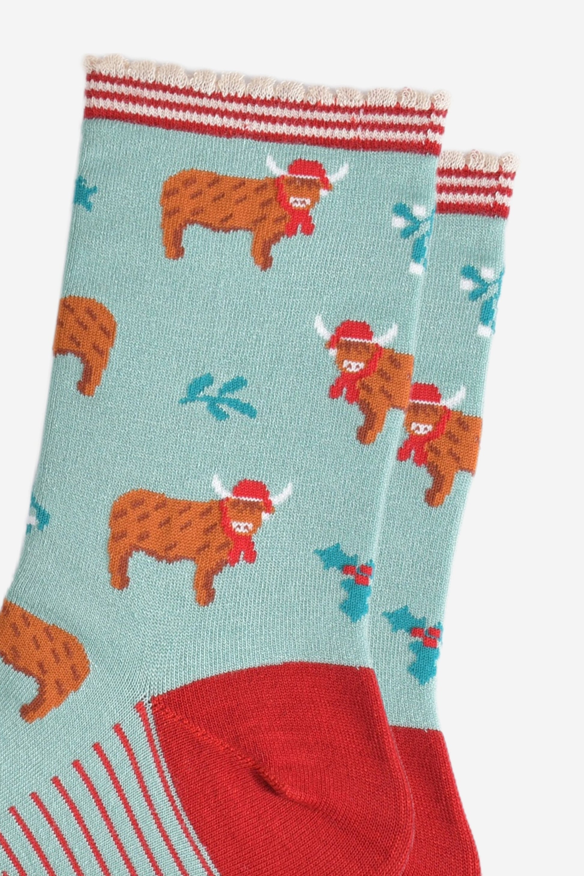 close up of the highland cow pattern on the socks, the cows are wearing red santa hats and red scarves, there are sprigs of holly and mistletoe throughout the design