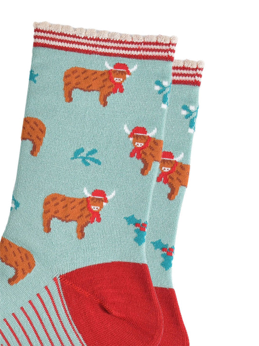 close up of the highland cow pattern on the socks, the cows are wearing red santa hats and red scarves, there are sprigs of holly and mistletoe throughout the design