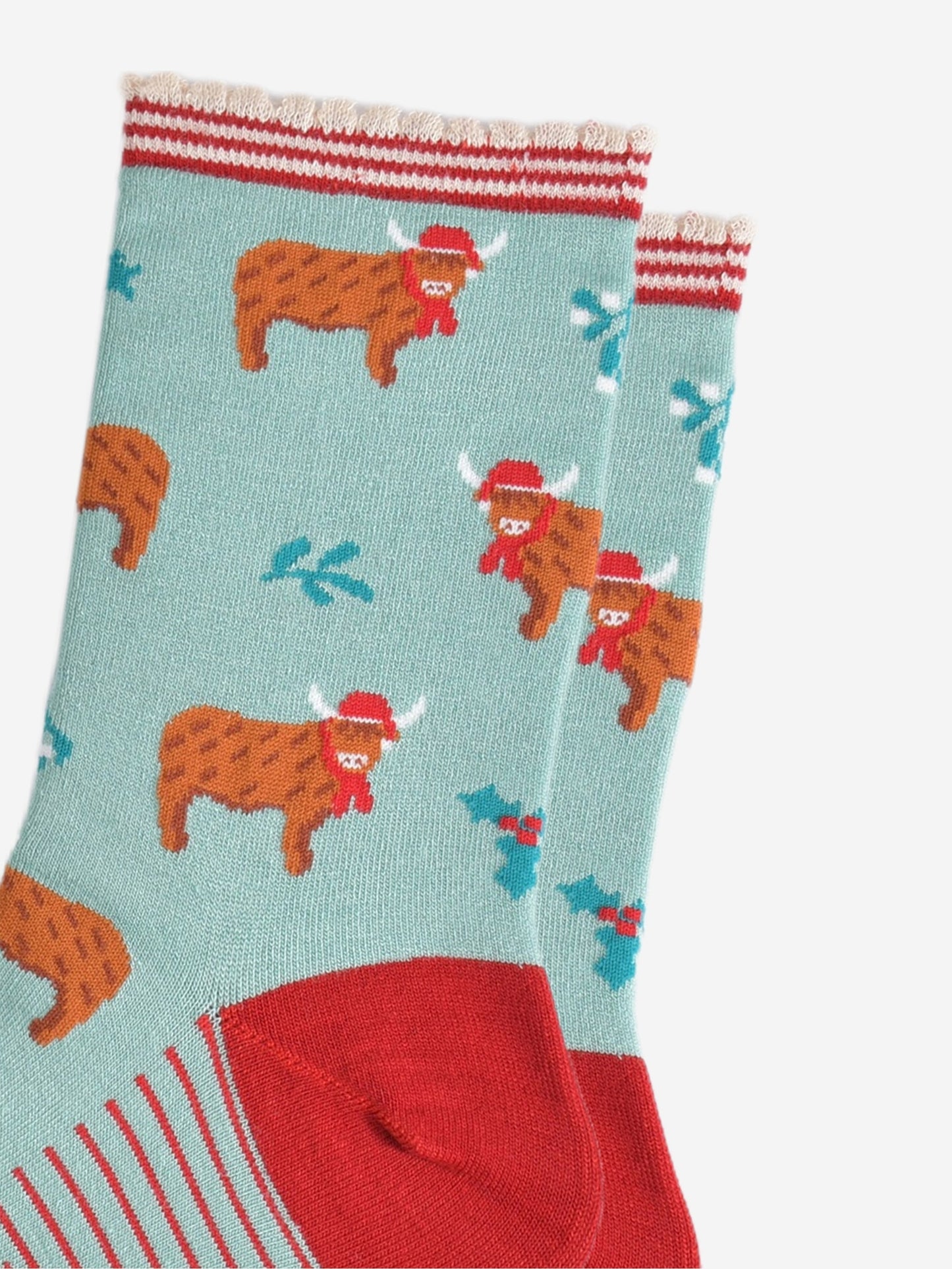 close up of the highland cow pattern on the socks, the cows are wearing red santa hats and red scarves, there are sprigs of holly and mistletoe throughout the design