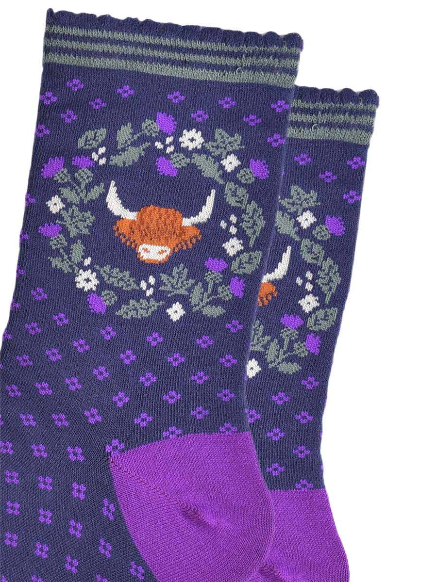 close up of the highland cow and wreath pattern on the ankle of the socks. the wreath is made from green and purple scottish thistles, the cow's head is in the middle of the wreath, the cow has long shaggy hair covering its eyes