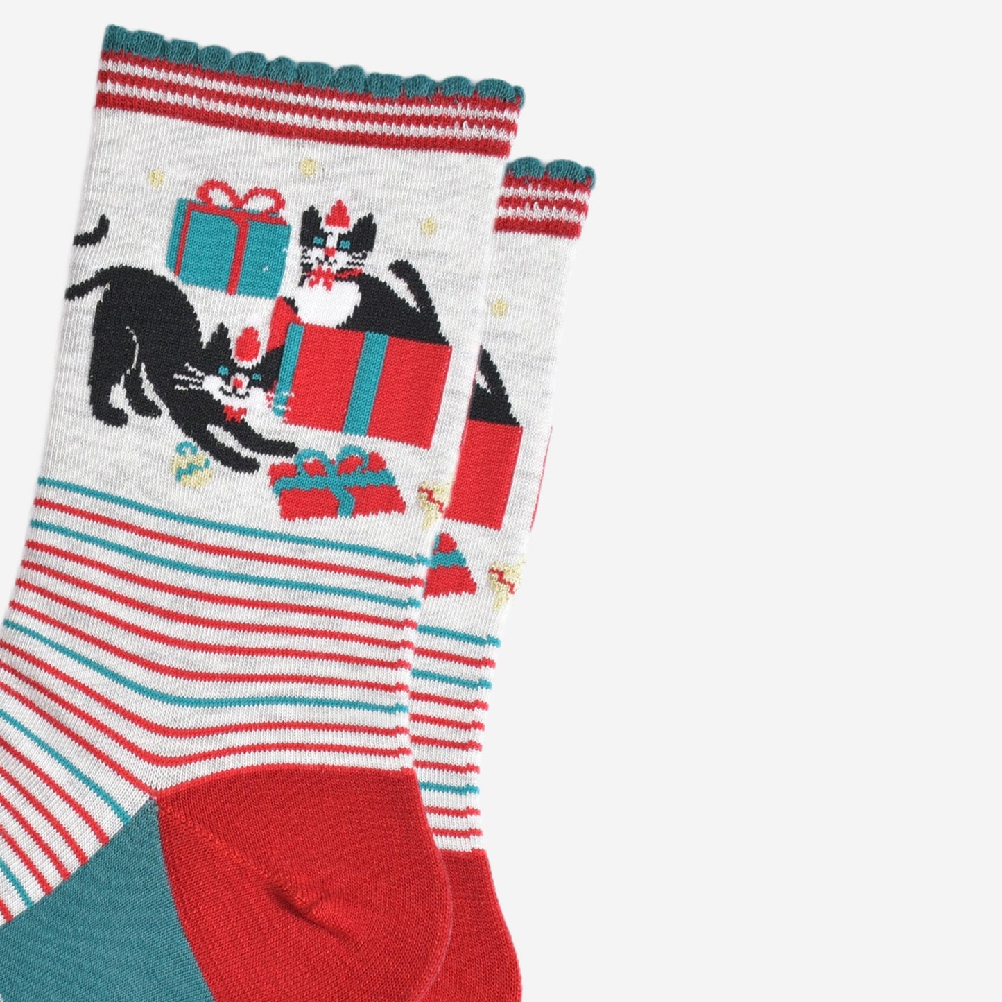 close up of the cats and christmas present pattern on the ankle of the socks, there is one cat sitting in a gift box, the other is playfully stretching while pawing at another wrapped present. both cats are black with white faces and are both wearing red santa hats and red bow ties