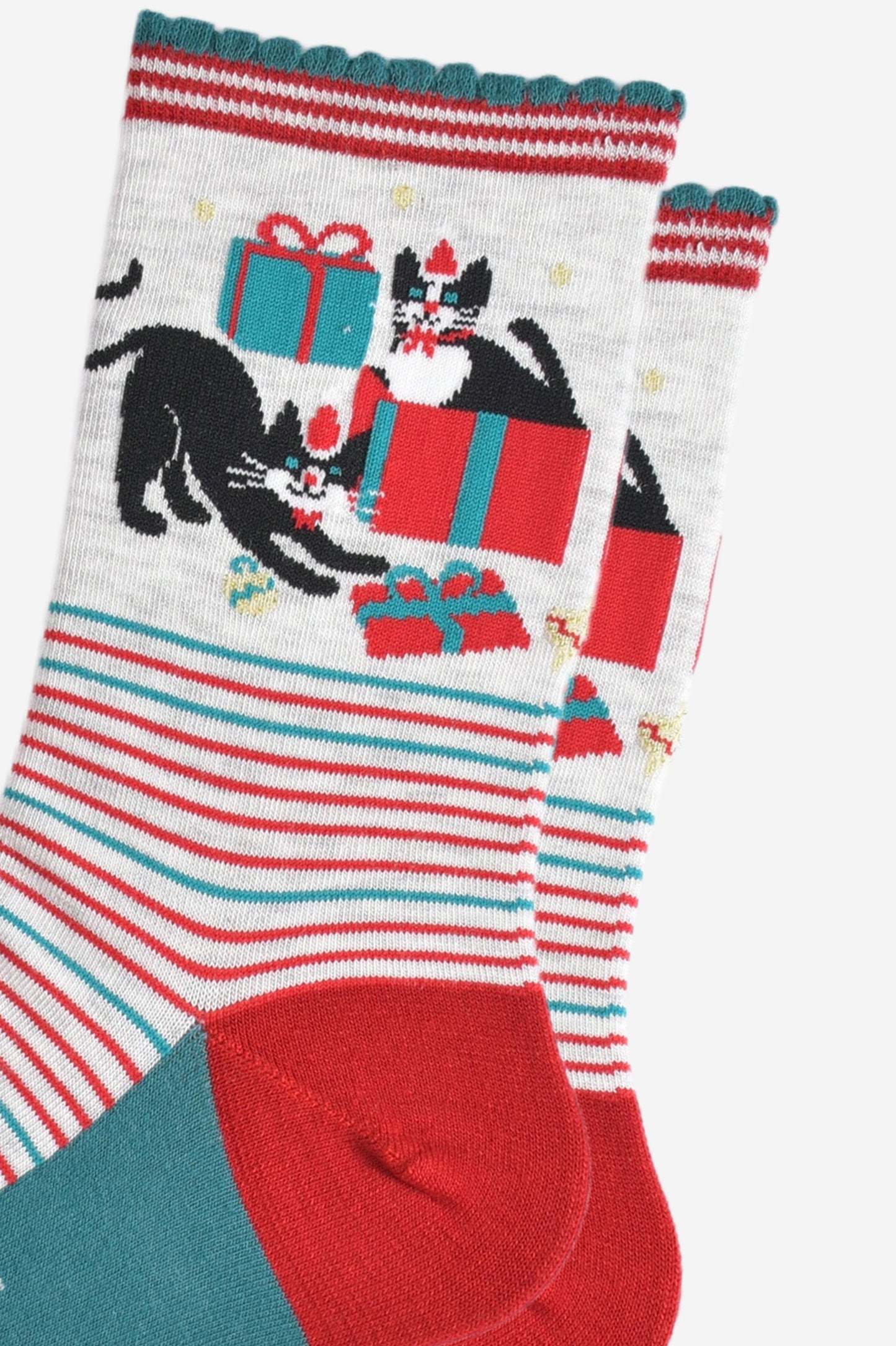 close up of the cats and christmas present pattern on the ankle of the socks, there is one cat sitting in a gift box, the other is playfully stretching while pawing at another wrapped present. both cats are black with white faces and are both wearing red santa hats and red bow ties