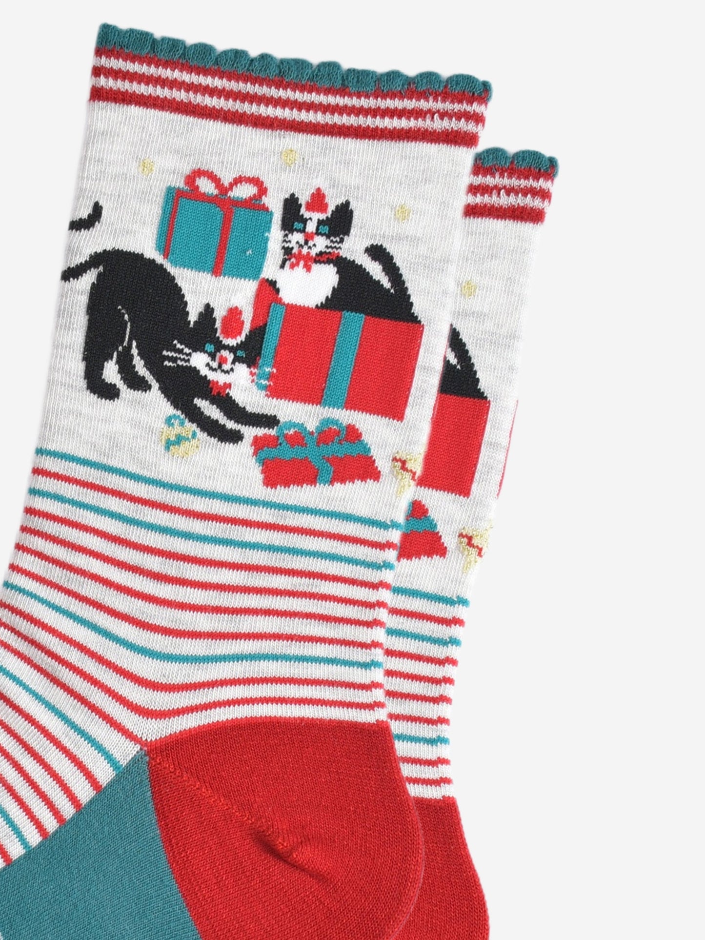 close up of the cats and christmas present pattern on the ankle of the socks, there is one cat sitting in a gift box, the other is playfully stretching while pawing at another wrapped present. both cats are black with white faces and are both wearing red santa hats and red bow ties