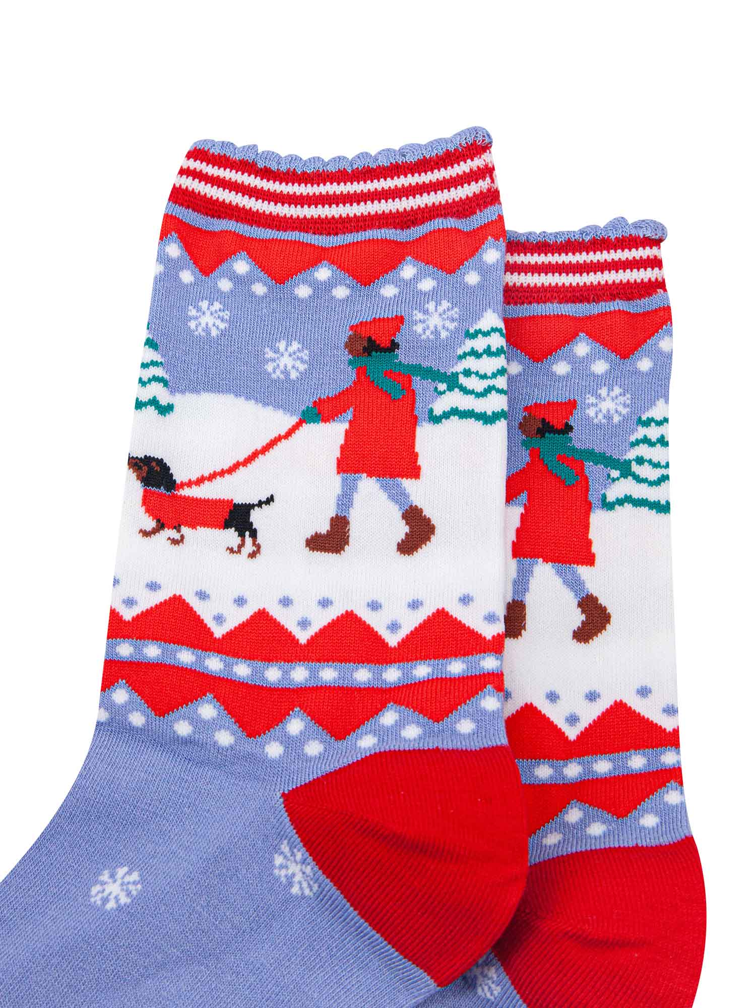 close up of the sausage dog and owner walking in the snow. both are wearing red winter coats, there are snow covered christmas trees in the background.