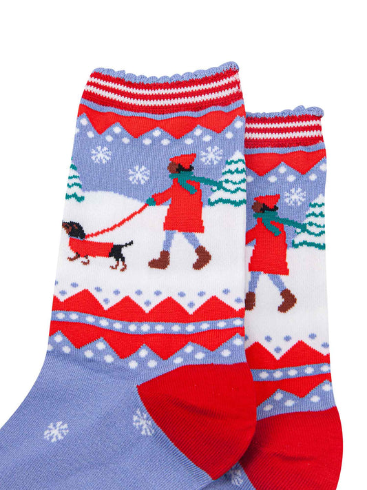 close up of the sausage dog and owner walking in the snow. both are wearing red winter coats, there are snow covered christmas trees in the background.