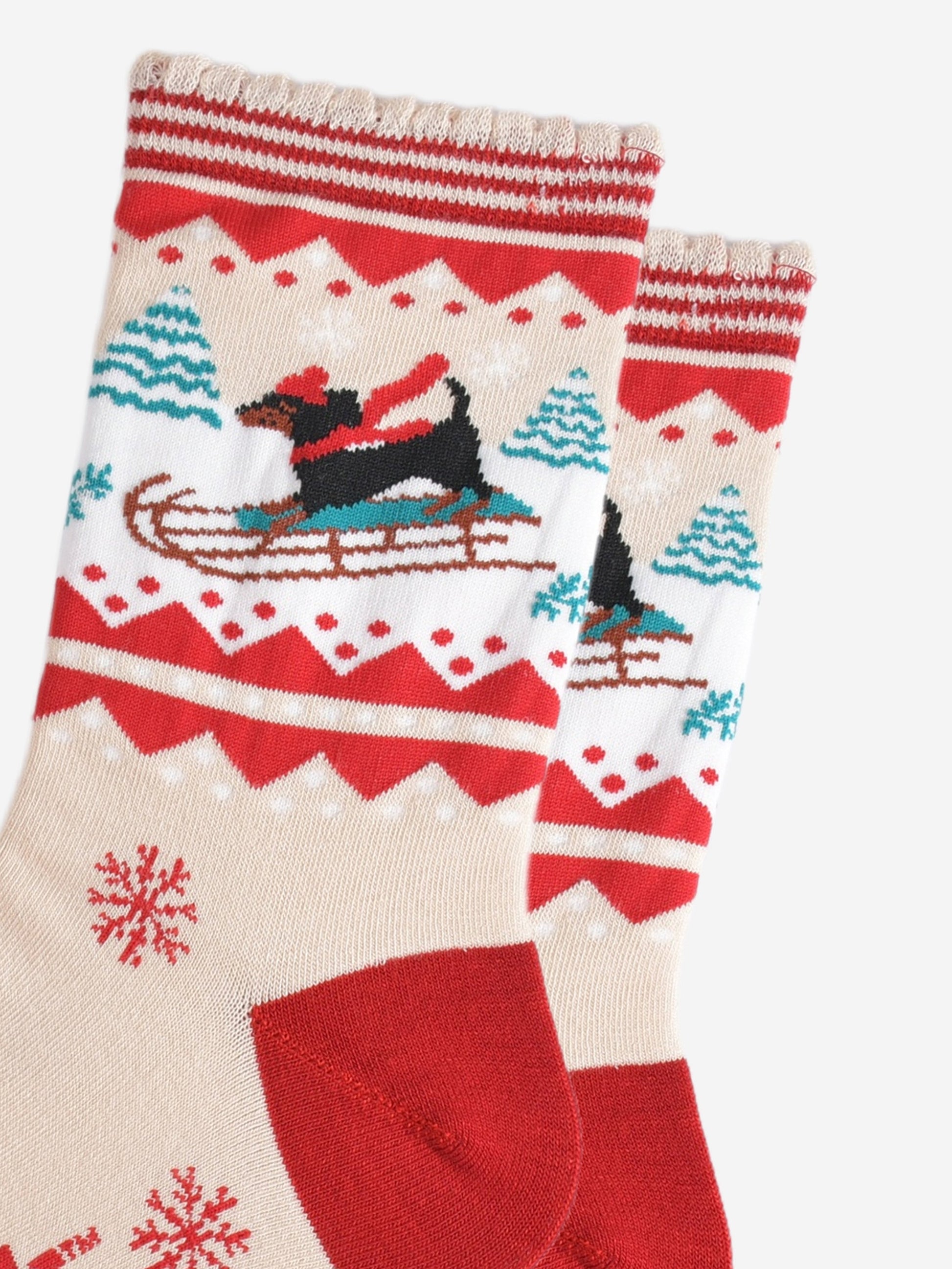 close up of the sledging sausage dog pattern, the dog is a traditional black dachshund, it is standing on a sledge and wearing a red winter hat and scarf, behind the dog are snow covered trees