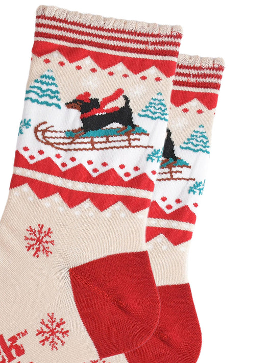 close up of the sledging sausage dog pattern, the dog is a traditional black dachshund, it is standing on a sledge and wearing a red winter hat and scarf, behind the dog are snow covered trees