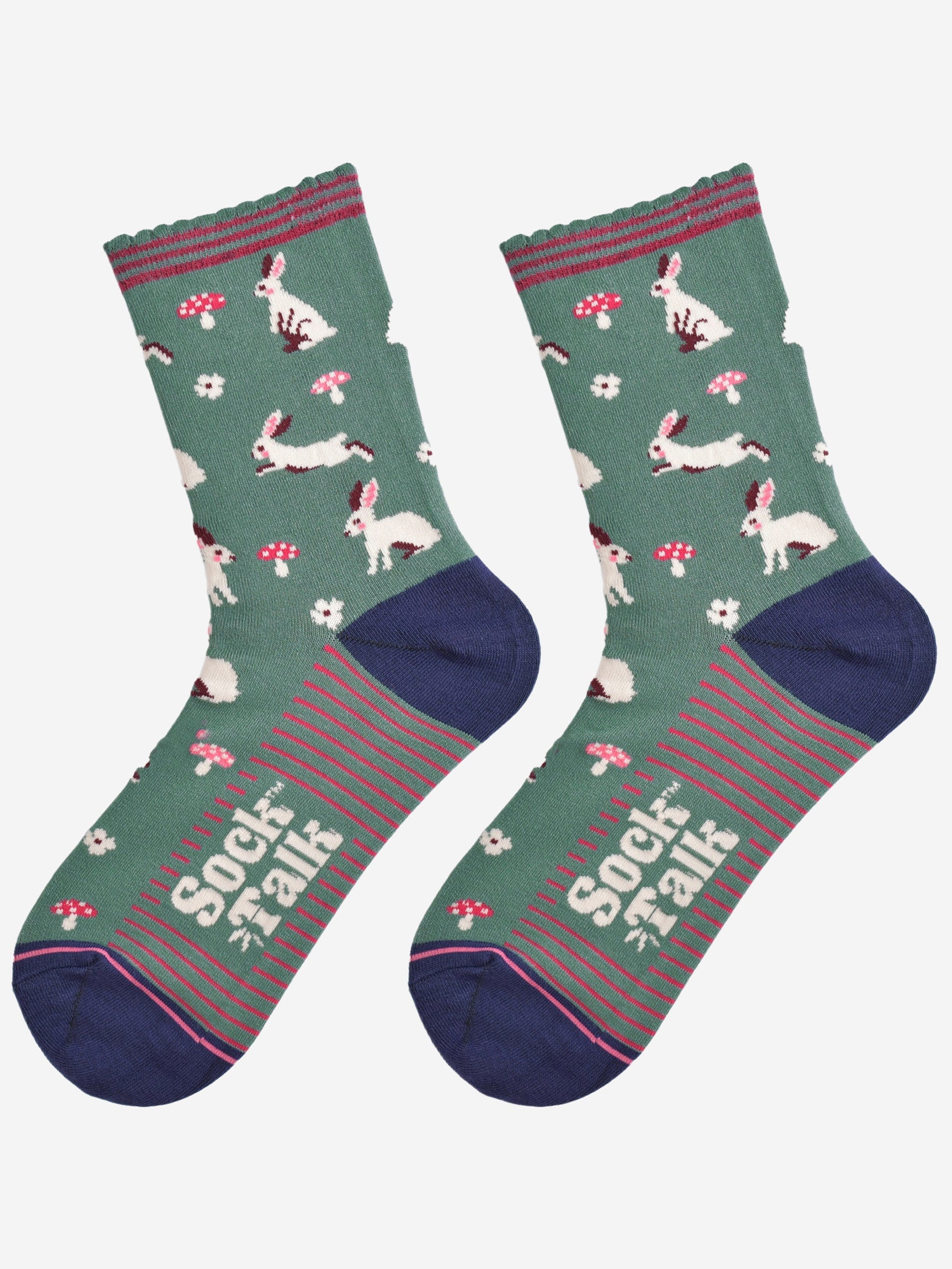 showing the woodland rabbit socks laying flat, there are white rabbits, hopping, sitting and standing among pink toadstools and white flowers, the solve of the socks has pink stripes in contrast to the green base of the design