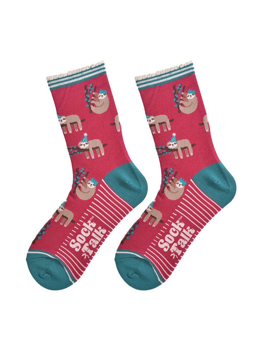 showing the festive sloth socks laying flat displaying the full pattern, there are sloths laying and climbing on tree branches while wearing green party hats, the sole of the socks have a white striped pattern