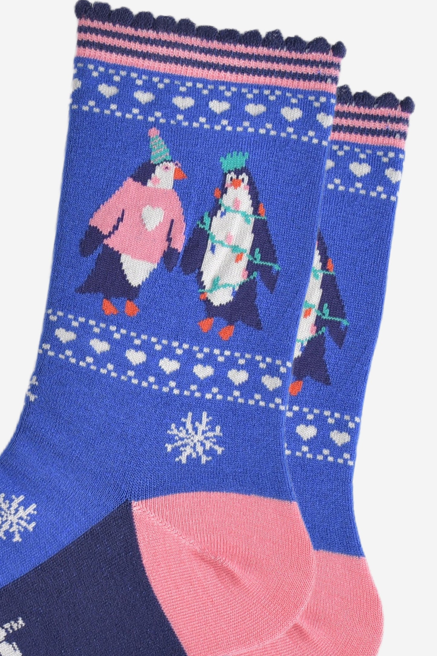 close up of the penguins on the ankle of the socks, one is wearing a pink xmas jumper and green party hat. the other penguin is entangled in festive party lights and is also wearing a crown shaped party hat