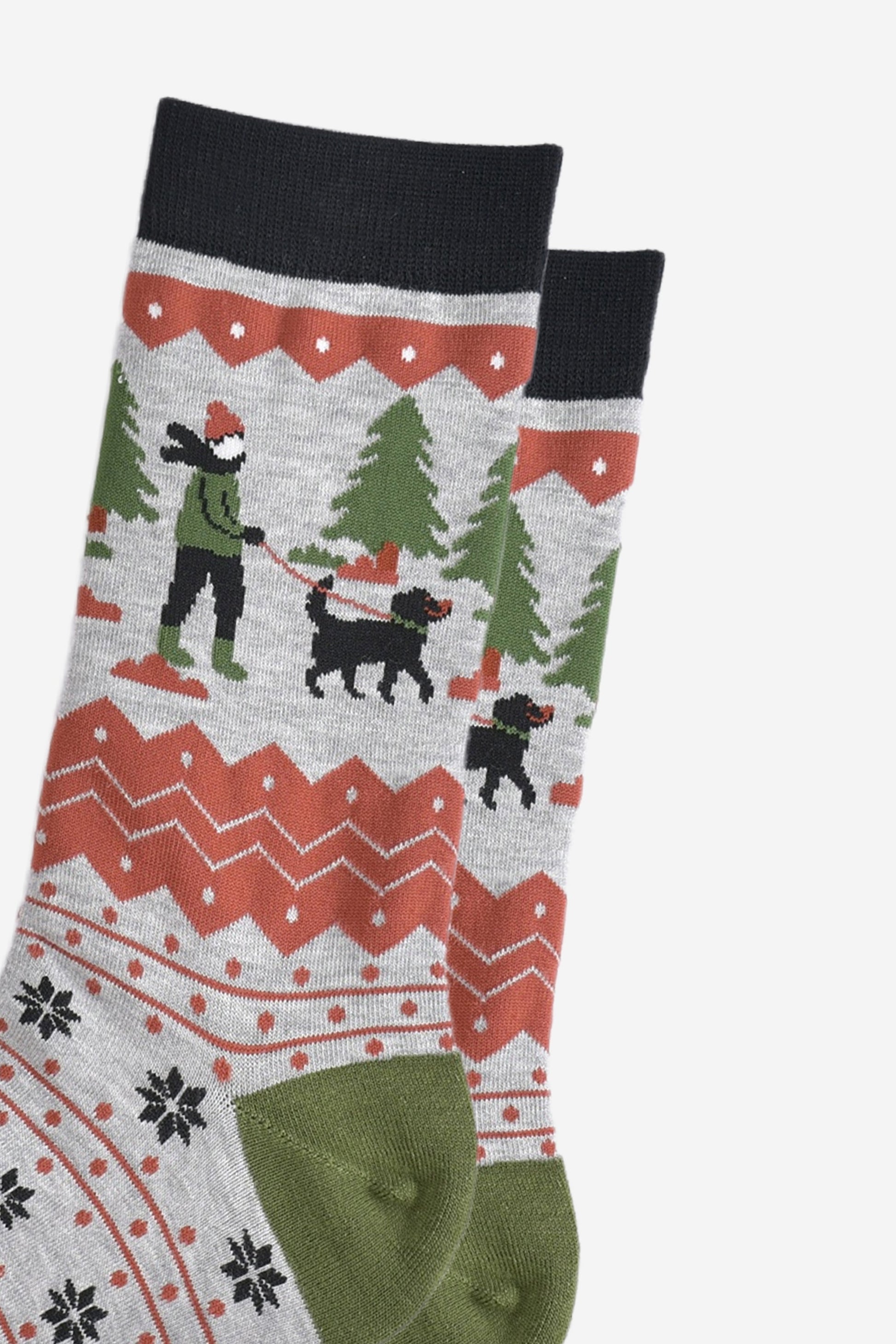 close up of the dog walker on the ankle of the socks. there is a black fog wit a green collar and red leash. the dog walker is wearing a green coat and boots, black trousers and coral pink winter hat in keeping with the colour tones of the socks