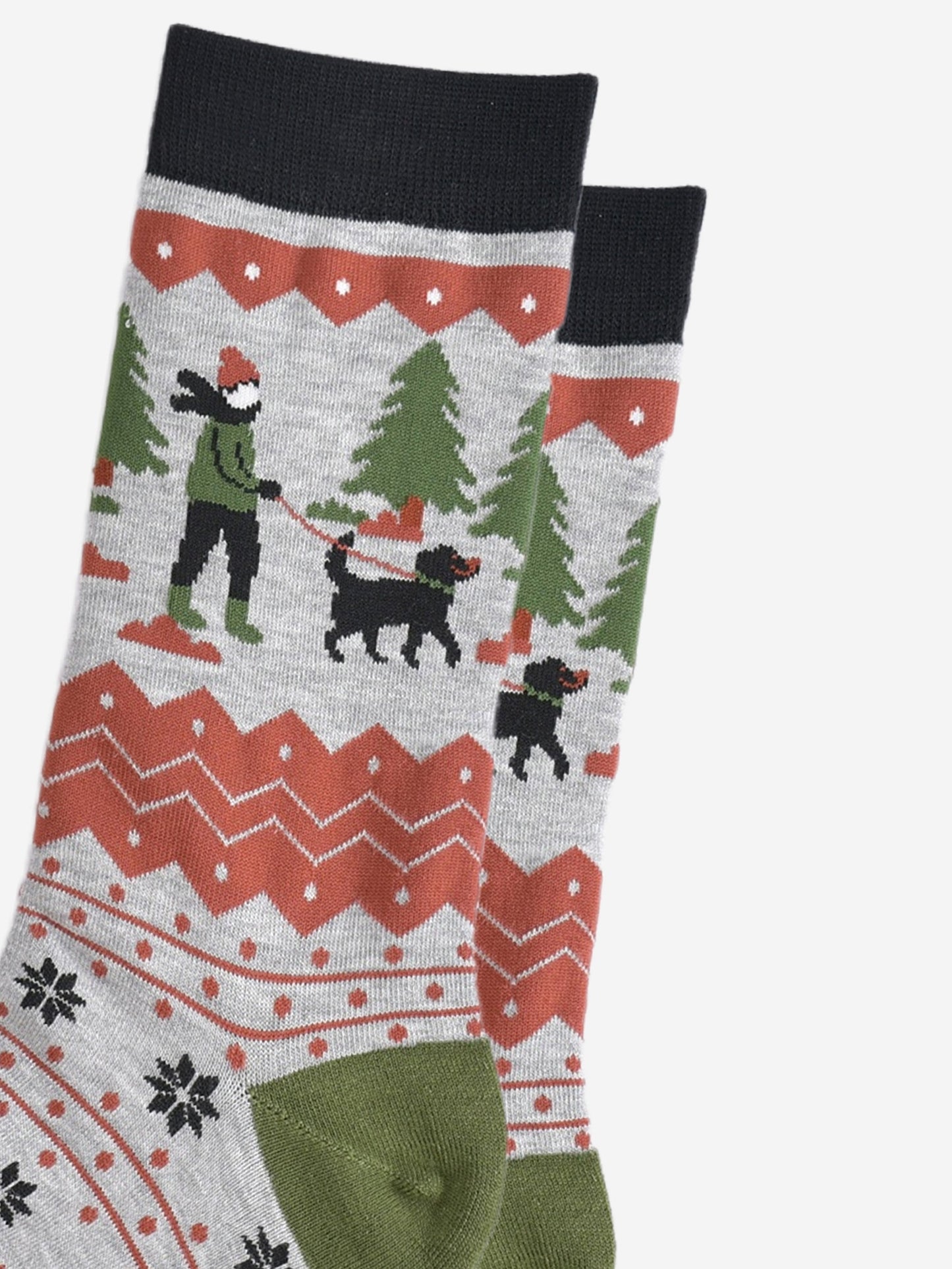 close up of the dog walker on the ankle of the socks. there is a black fog wit a green collar and red leash. the dog walker is wearing a green coat and boots, black trousers and coral pink winter hat in keeping with the colour tones of the socks