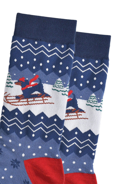 close up of the sledging sausage dog on the ankle of the socks, the dog is wearing a red hat and scarf and there are snow covered christmas trees in the background

