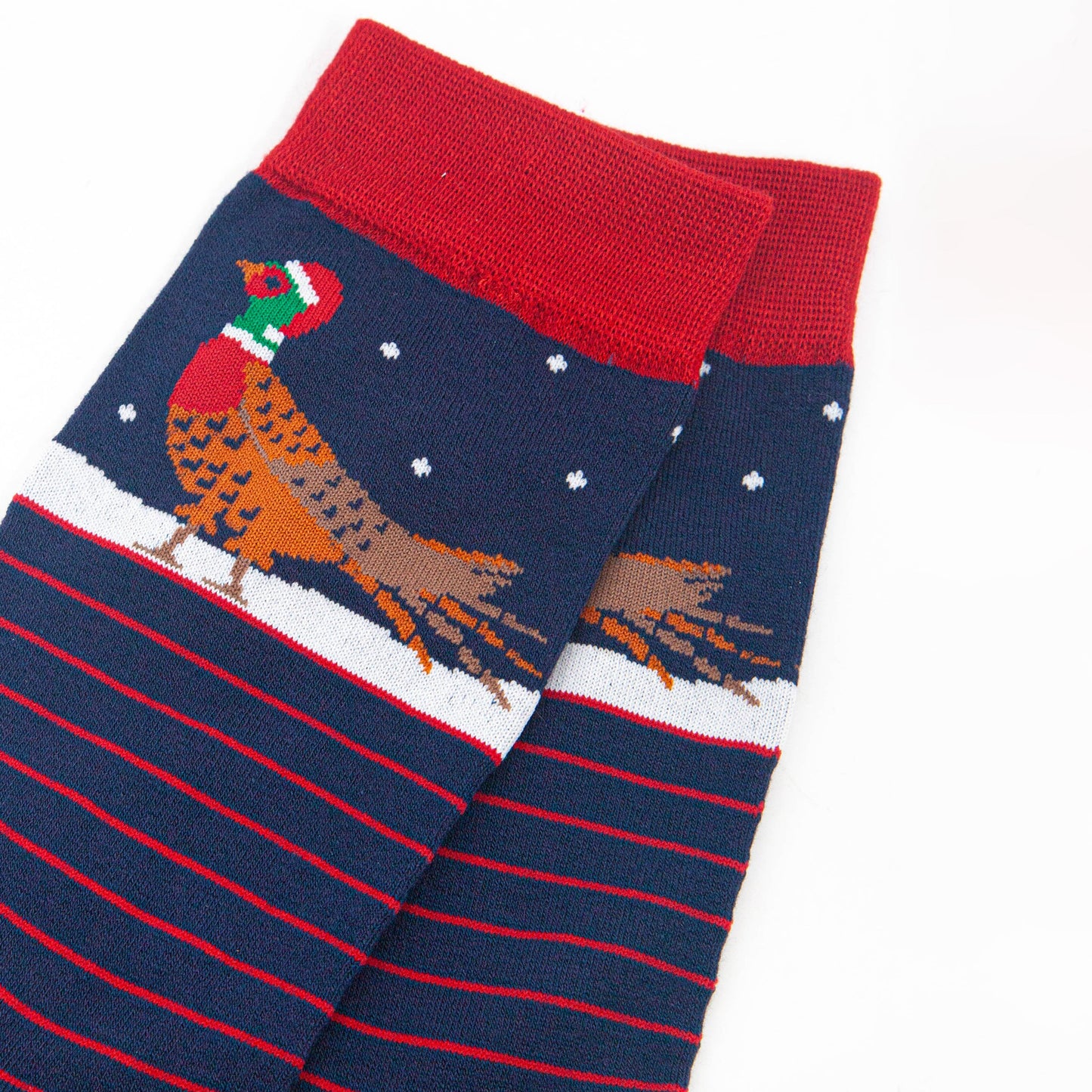 close up of the pheasant on the ankle of the socks, it is walking on snow and wearing a red santa hat, there are small white snowflakes falling around it