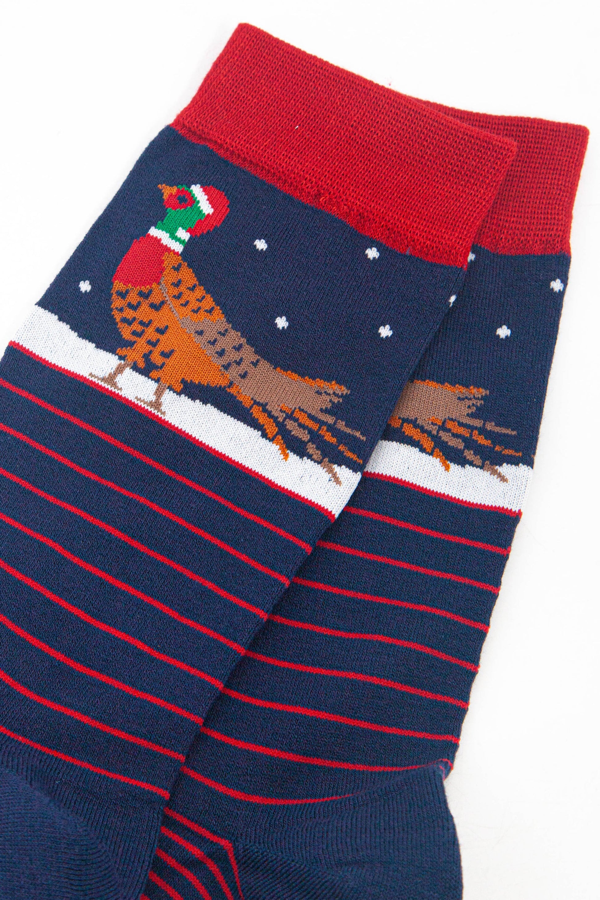 close up of the pheasant on the ankle of the socks, it is walking on snow and wearing a red santa hat, there are small white snowflakes falling around it